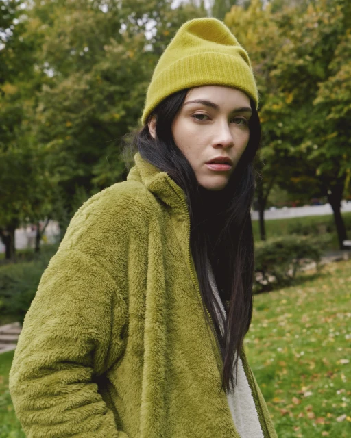 Portrait of a young woman in a green fuzzy jacket and knit beanie, posing in a green outdoor park setting on a sunny day.