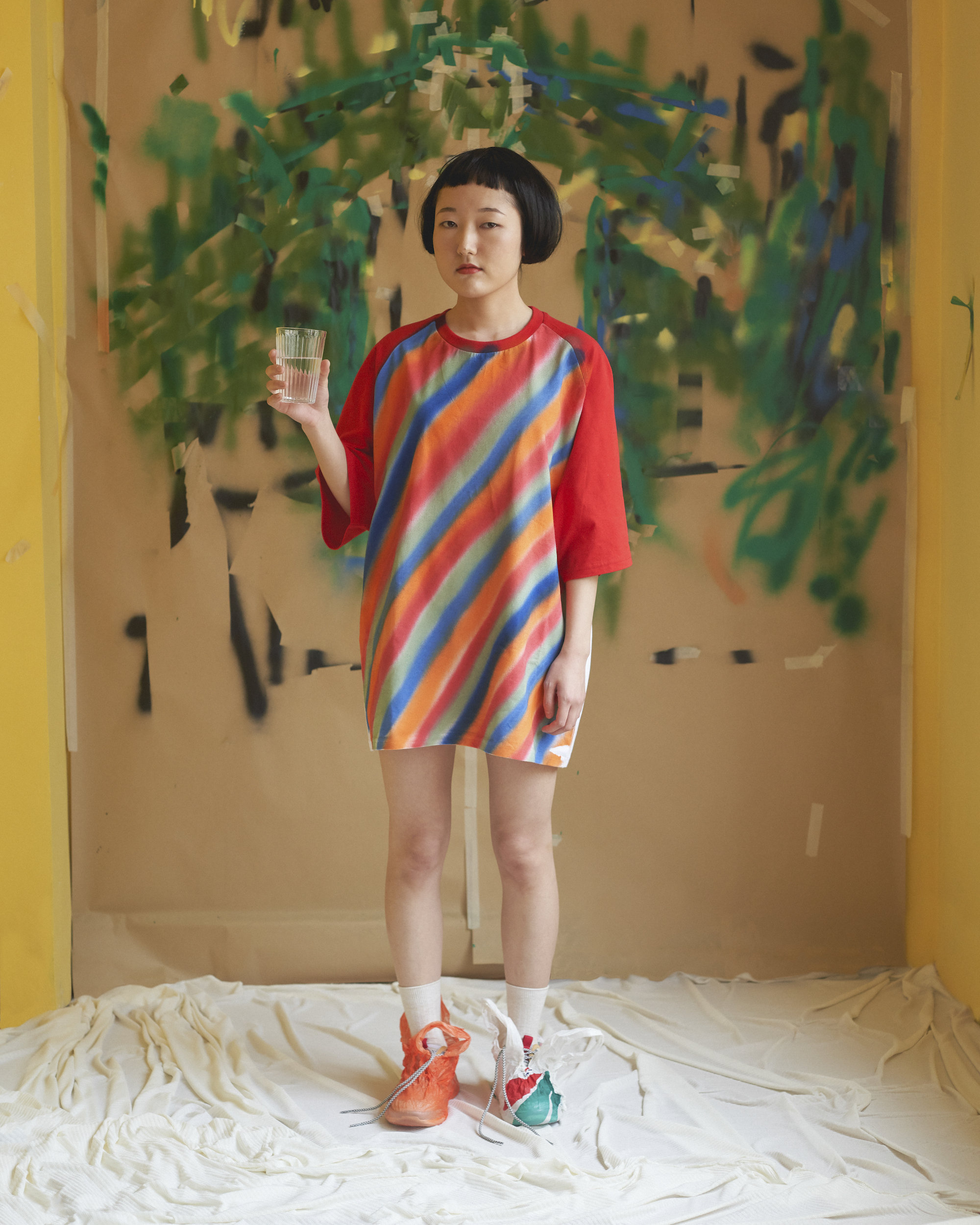 Full shot of a young woman with short black hair in colorful dress holding a glass of water in an abstract studio setting.