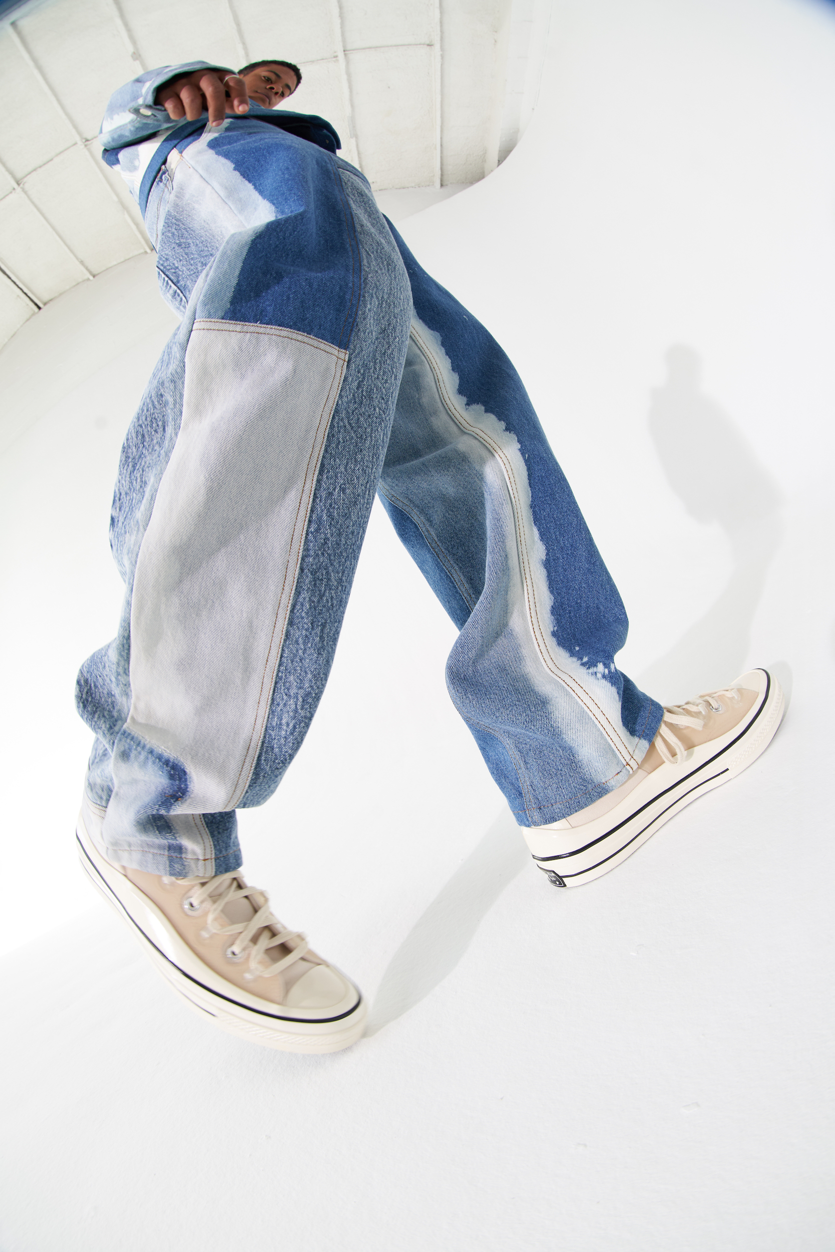 Close-up, low angle shot of a person wearing patchwork denim pants and beige sneakers, standing against a bright white background.