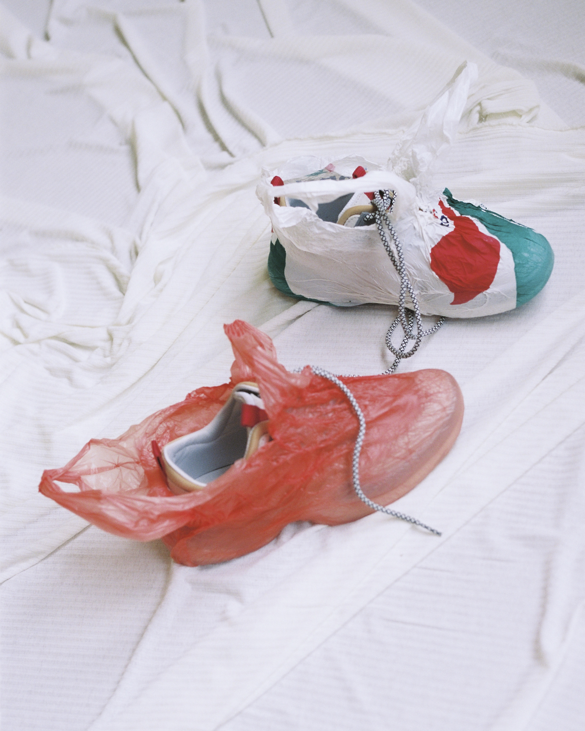 Two colorful painted sneakers encased in transparent plastic bags rest on a wrinkled white sheet in a softly lit indoor setting.