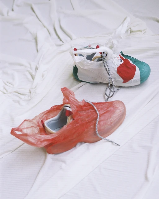 Two colorful painted sneakers encased in transparent plastic bags rest on a wrinkled white sheet in a softly lit indoor setting.