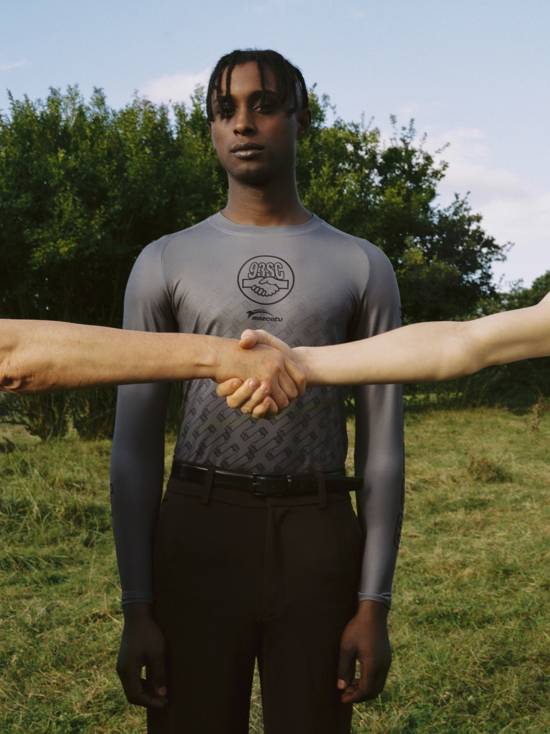 A light-skinned person shakes hands with two other people wearing a gray long-sleeve shirt against an outdoor grassy background.