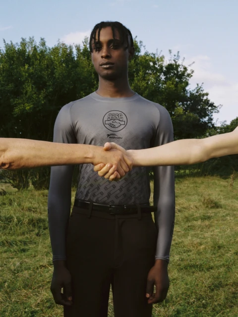 A light-skinned person shakes hands with two other people wearing a gray long-sleeve shirt against an outdoor grassy background.