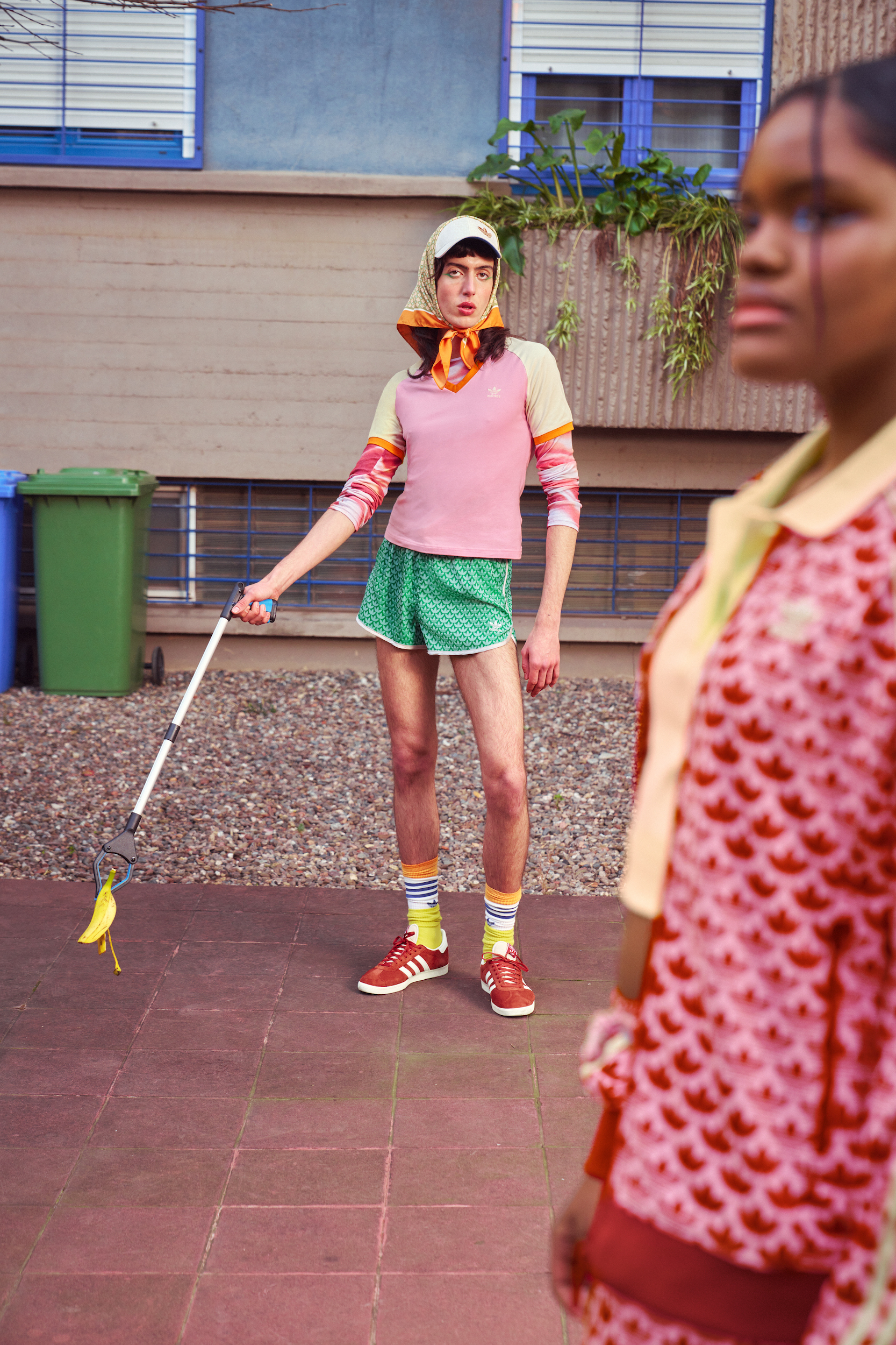 Two women in retro Adidas outfits pose outdoors; one holds a banana-peel-covered grabber against a building backdrop with trash bins and window plants.