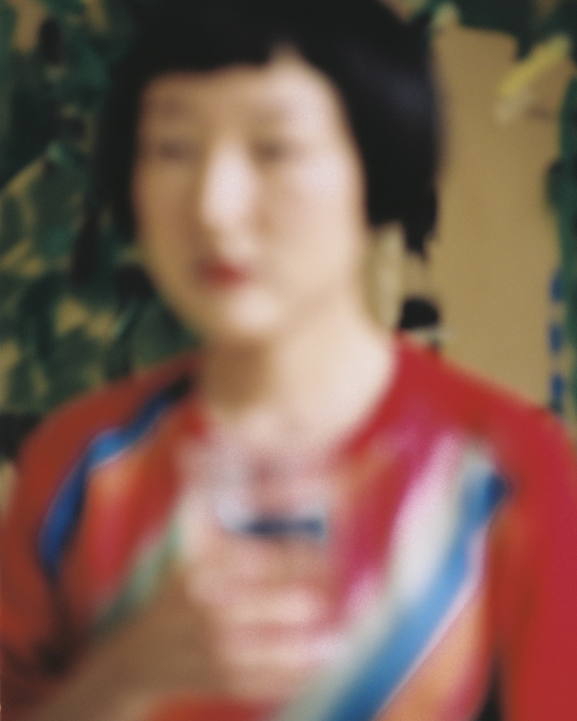 An out-of-focus shot of an Asian woman holding a glass in front of a leafy green background, wearing a red and blue top.