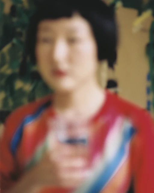 An out-of-focus shot of an Asian woman holding a glass in front of a leafy green background, wearing a red and blue top.