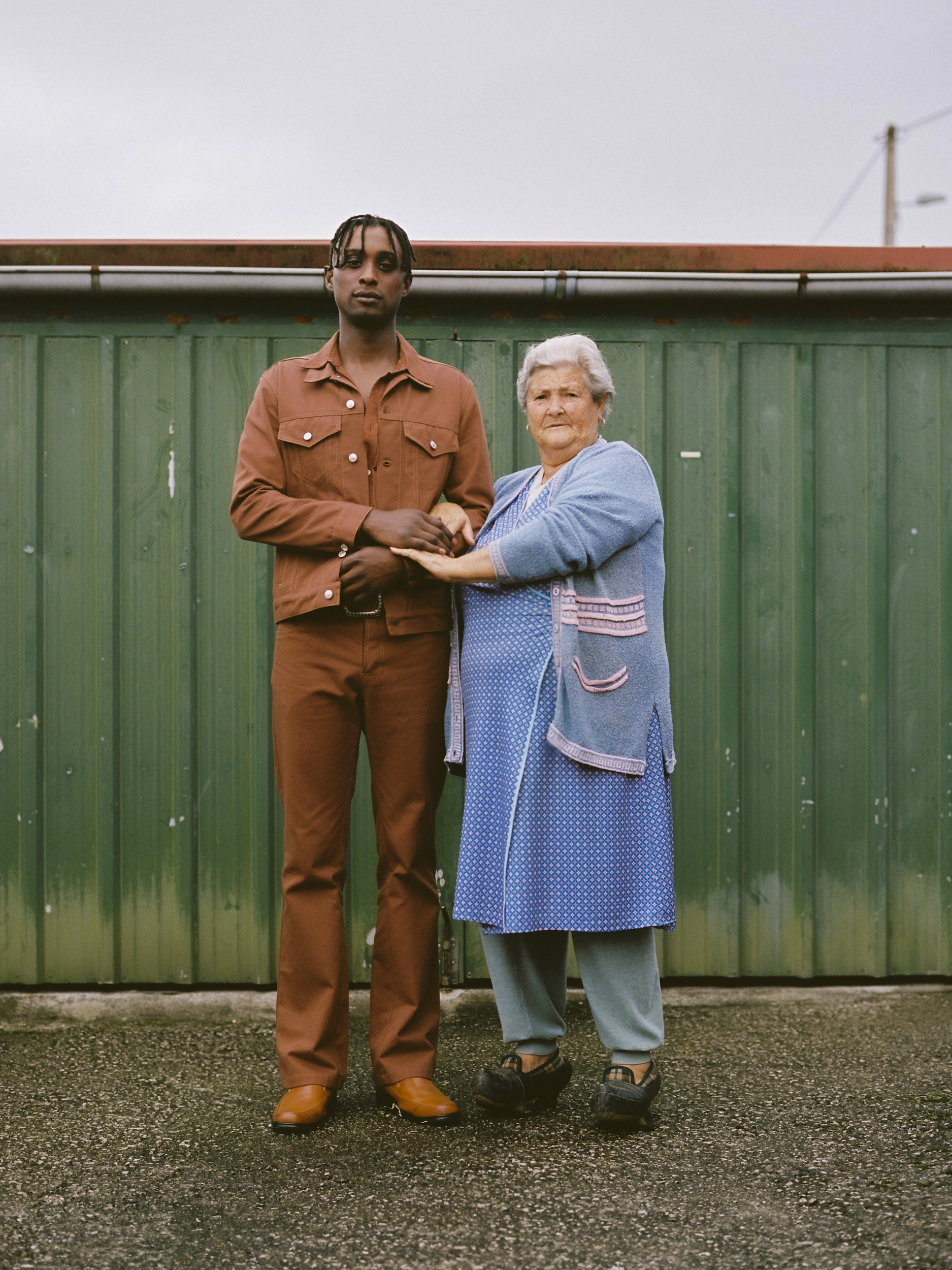Portrait of an adult man and an elderly woman standing close together in front of a green metal fence in an outdoor environment.