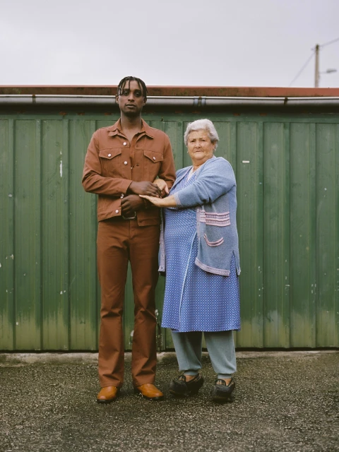 Portrait of an adult man and an elderly woman standing close together in front of a green metal fence in an outdoor environment.
