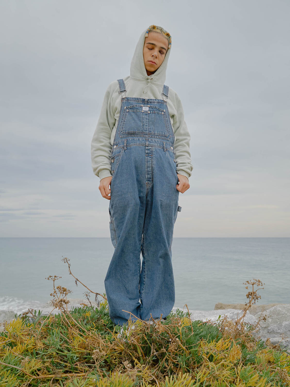 Young man wearing denim overalls and a grey hoodie standing near the ocean by Alexandra Cepeda