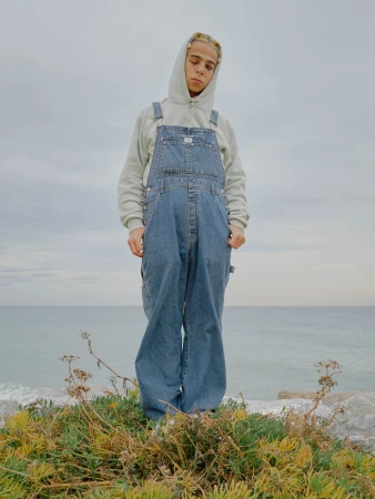 Young man wearing denim overalls and a grey hoodie standing near the ocean by Alexandra Cepeda