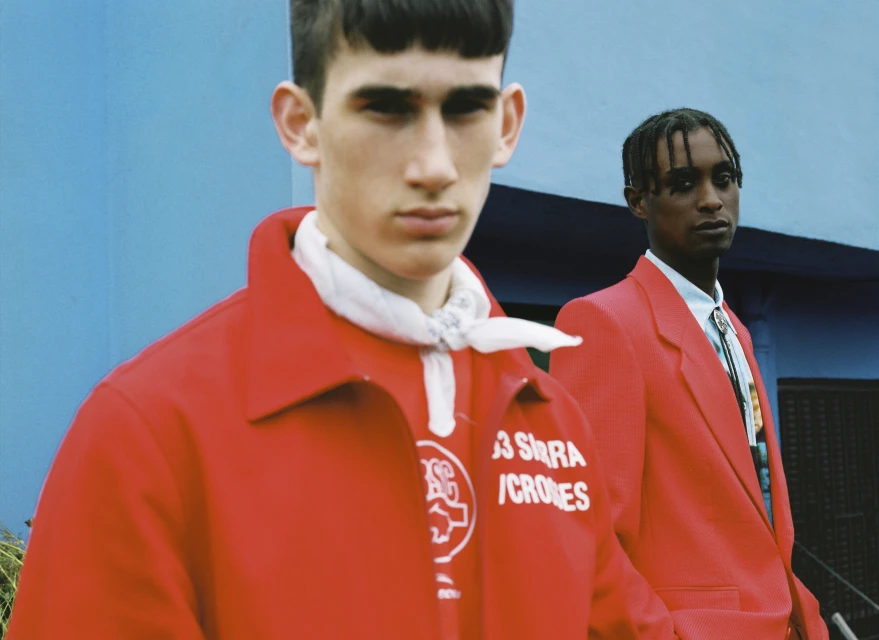 Two young adult males pose in red jackets against a blue wall, creating a striking fashion editorial shot with a vibrant and modern aesthetic.