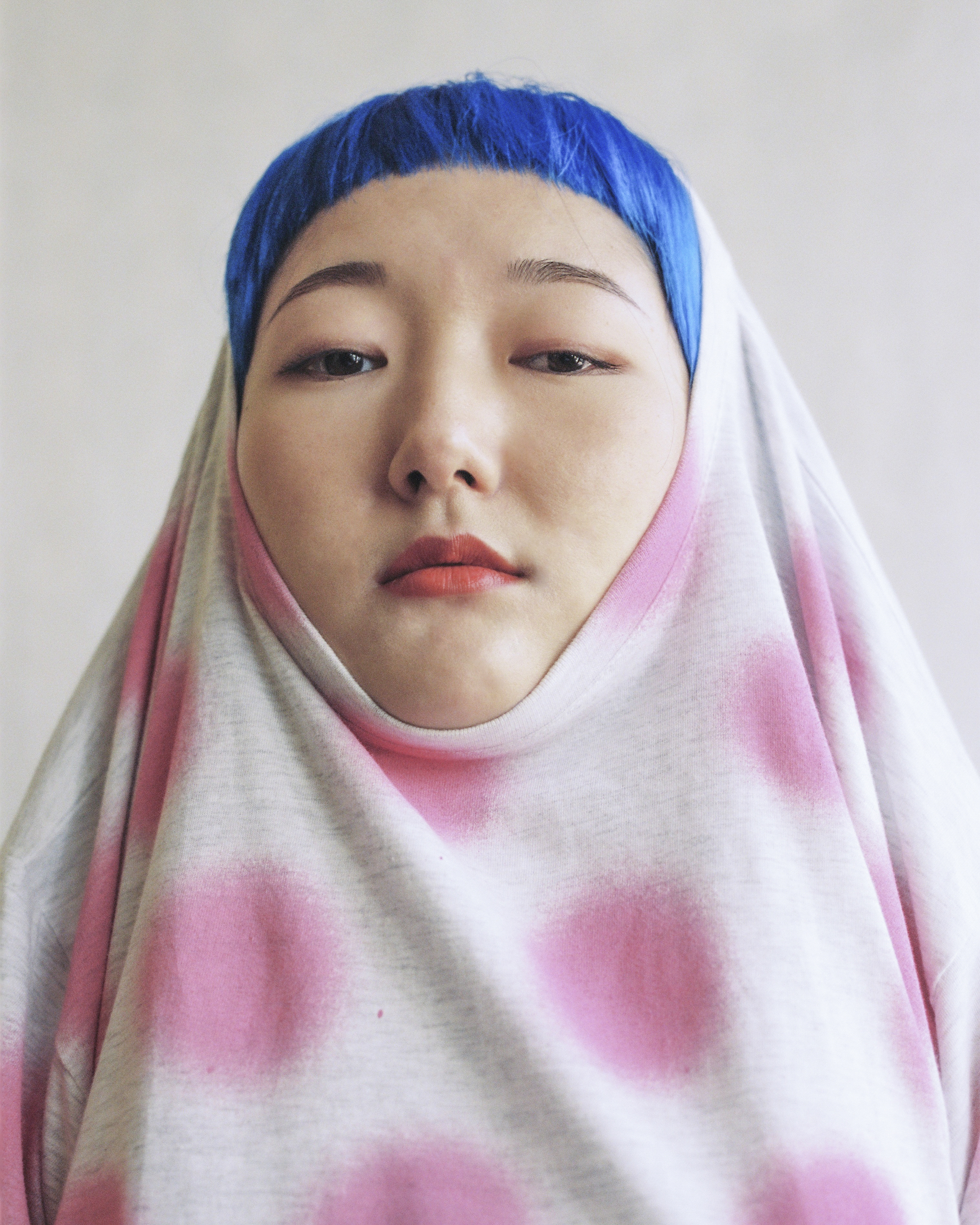 Portrait of an Asian woman with blue hair covered in a gray t-shirt with pink polka dots, looking directly at the camera against white.