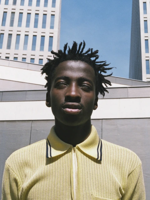 Portrait of a young Black man with dreadlocks wearing a yellow knit polo shirt, standing outdoors against a modern architectural backdrop on a sunny day.