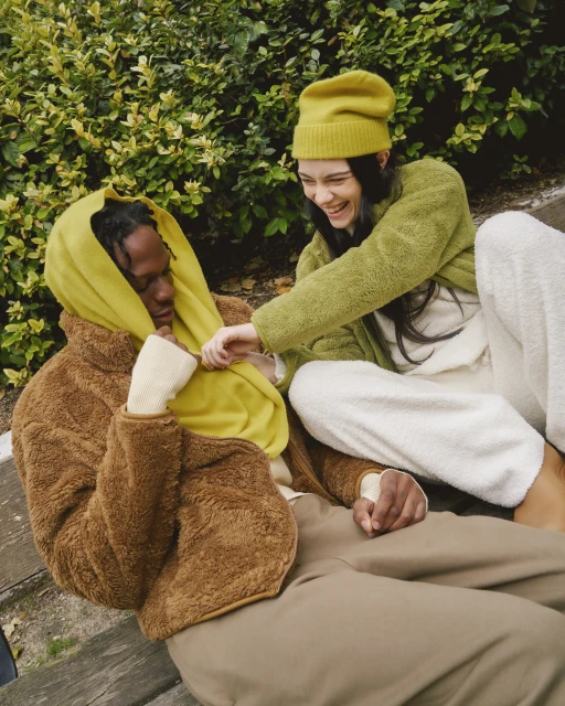 A happy couple relaxes on a bench outdoors; the woman, with a beanie and green jacket, playfully adjusts the yellow scarf of the man.