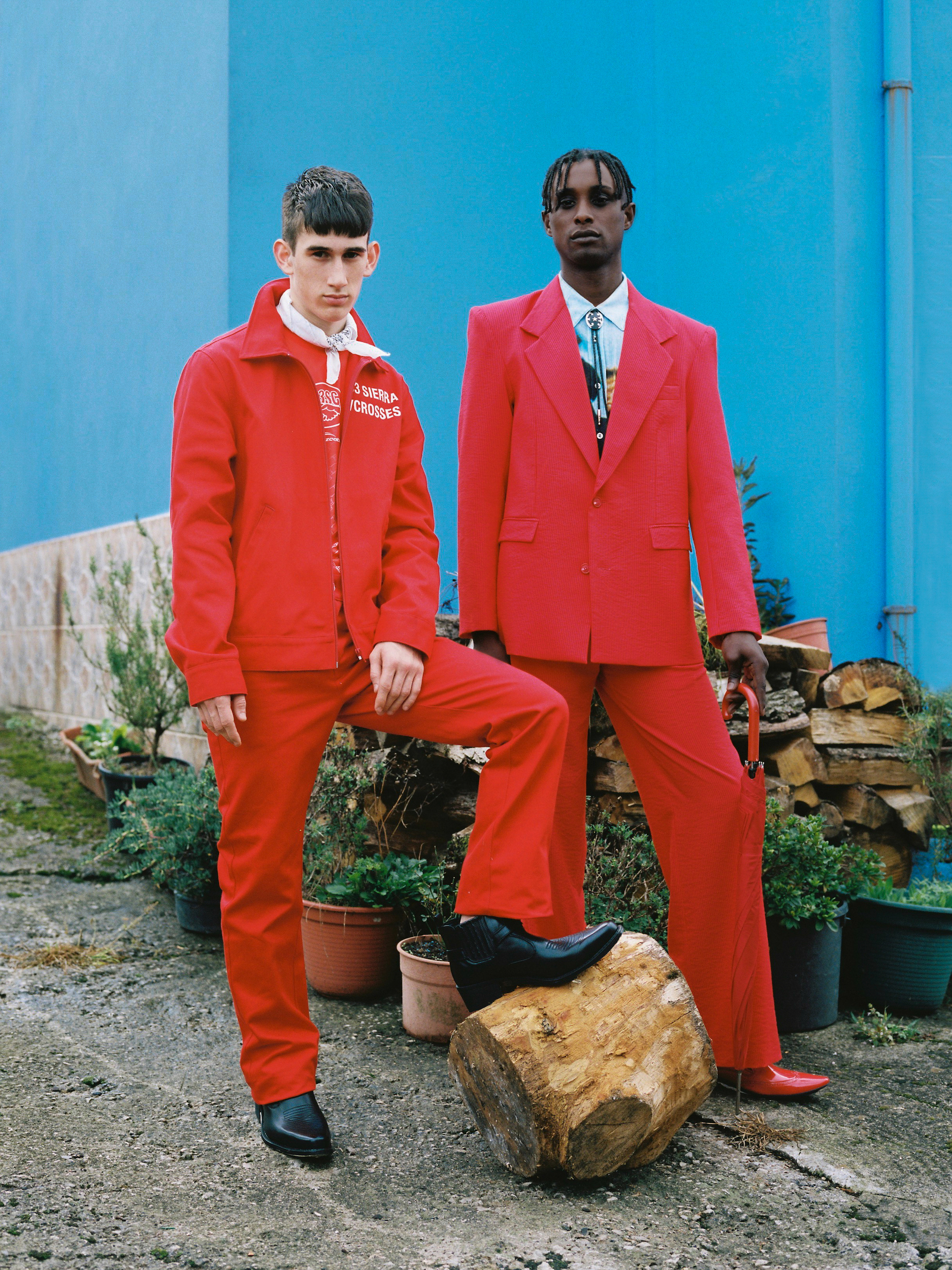 Two men in bright red suits posing outdoors against a vibrant blue wall with potted plants and a wood log foreground.