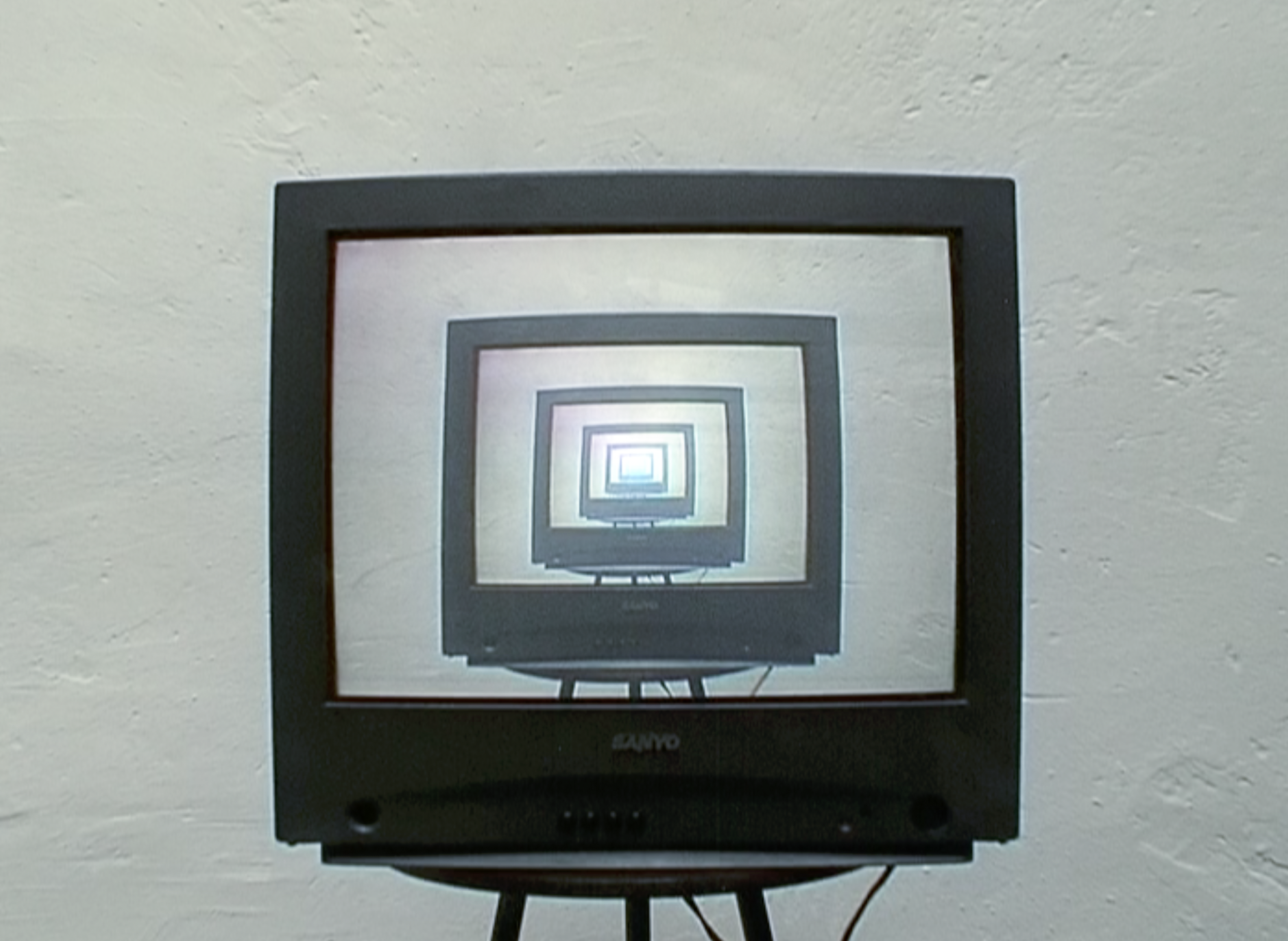 A black cathode-ray television on a stand shows a recursive screen display, diminishing into the distance, against a white wall.