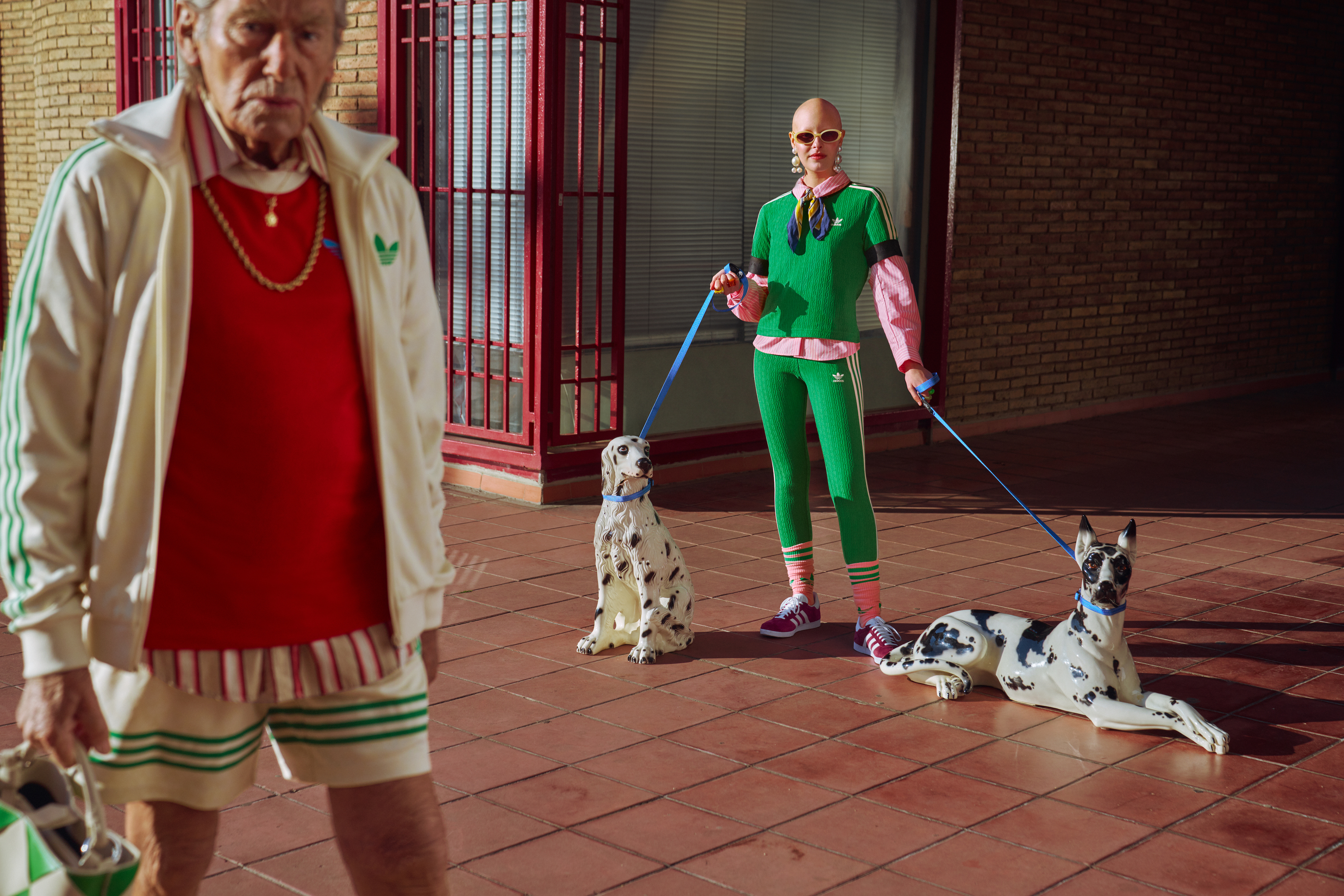 An older and younger adult wearing retro Adidas tracksuits pose outside with leashed Dalmatians, blending fashion and pet companionship.
