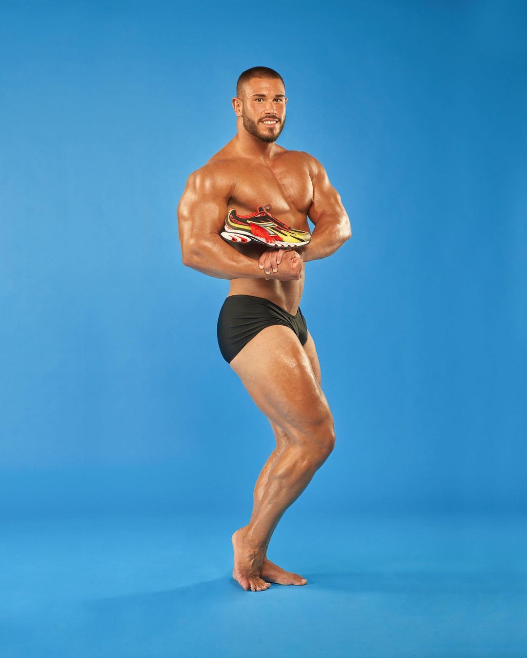 Athletic male flexing with running shoe in a photo studio. Man is tanned and posing with a barefoot stance and wearing black trunks.
