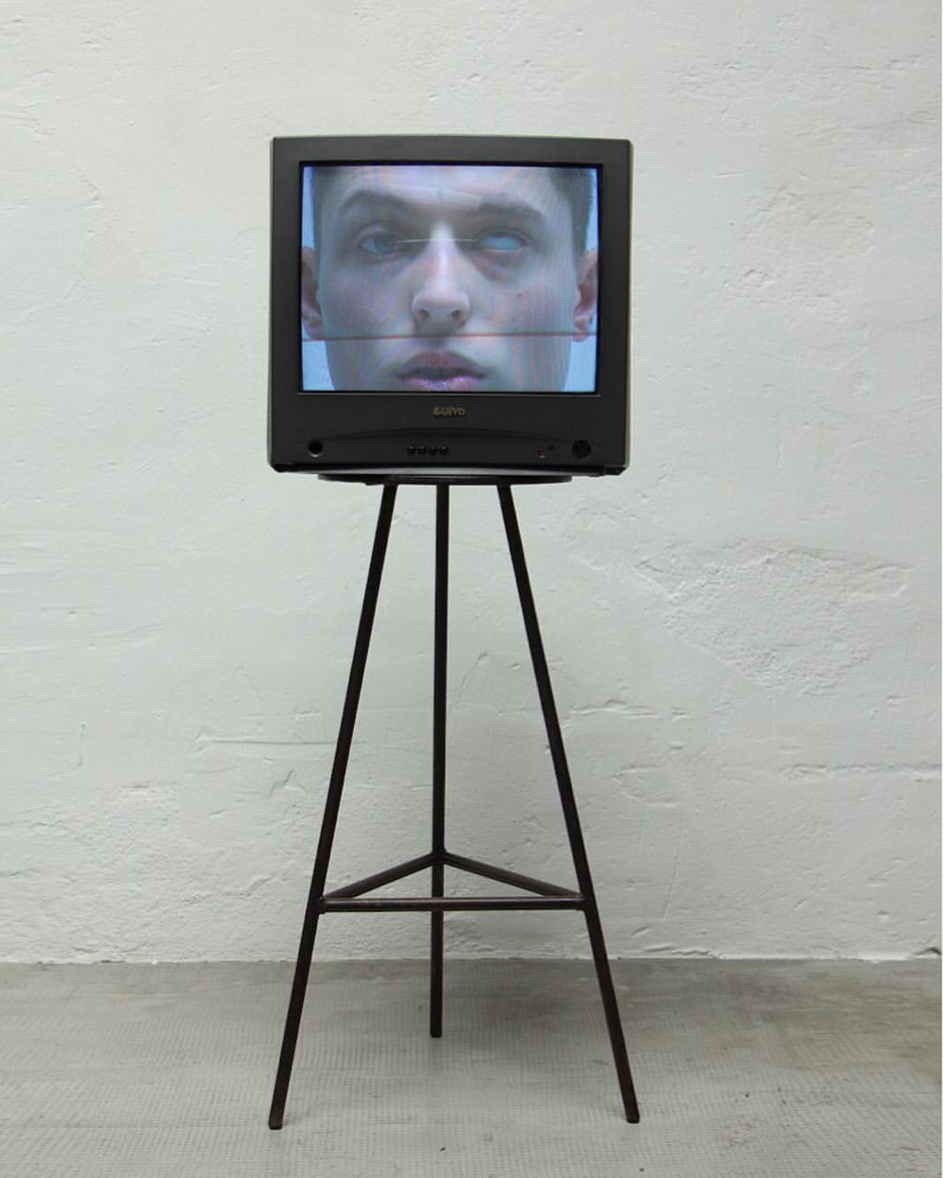 Vintage television on tripod stand displaying a man's face with distorted lines. The artwork is set against a textured white wall.