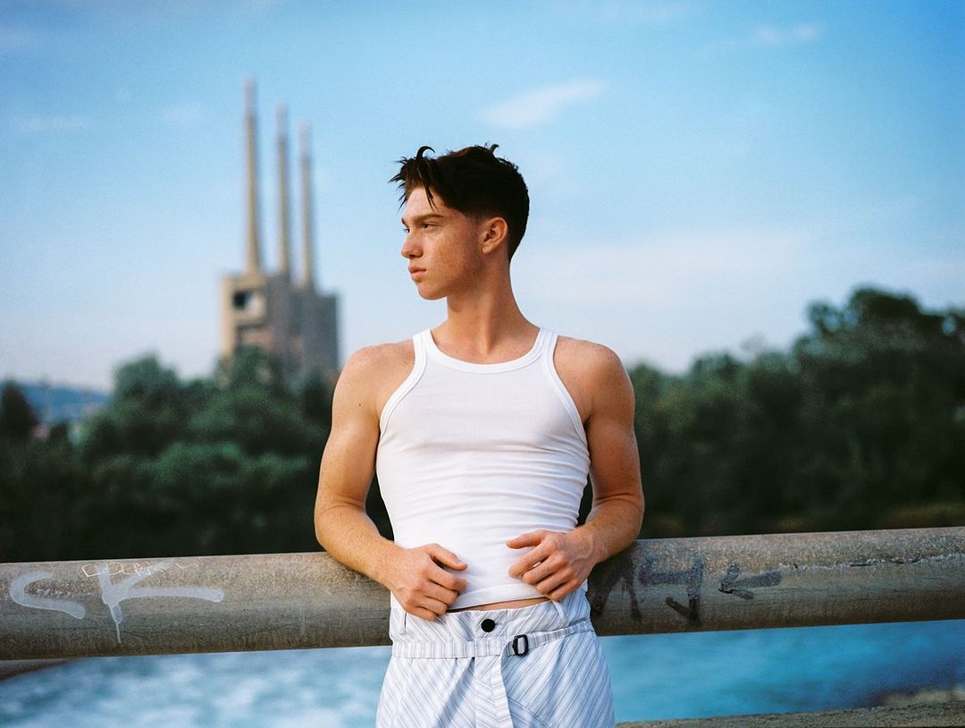 Young adult male in white tank top and striped pants leans against a metal railing with industrial buildings and green trees in background.