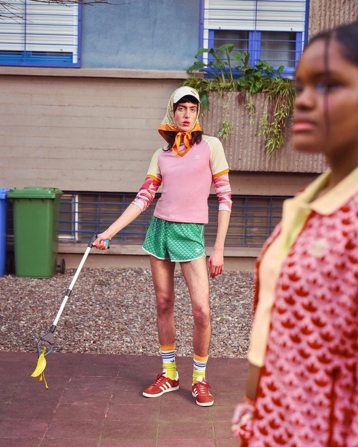 Model wearing a headscarf, pink top, and green shorts holding a litter picker outdoors with another person visible in the foreground