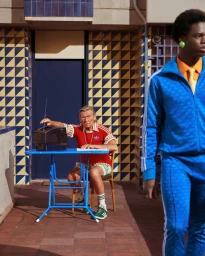 An elderly man in a red top adjusting a radio at a blue table, with a young Black man in a blue tracksuit in the foreground.