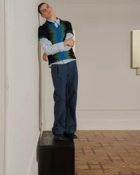 Young male model leaning against an off-white wall wearing blue trousers, a light blue shirt, and a black patterned vest.