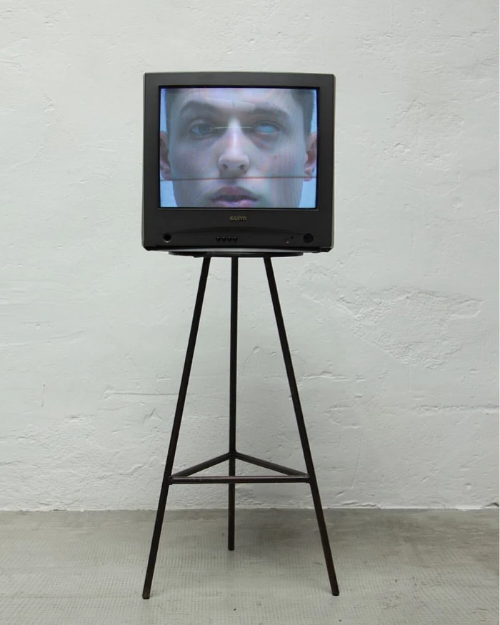Vintage television displaying a man's face on screen, resting on a tall metal tripod against a textured grey wall.