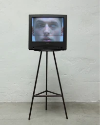 Vintage television displaying a man's face on screen, resting on a tall metal tripod against a textured grey wall.