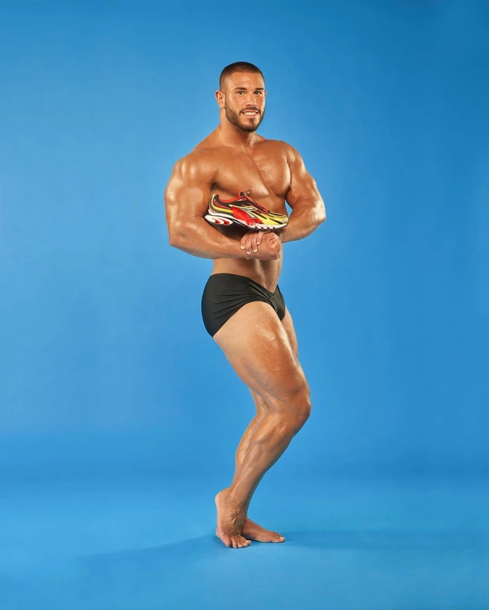Muscular man posing against a blue seamless background holding a colourful sports sneaker against his chest