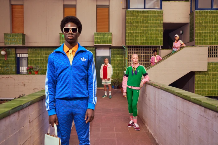 Black man in blue tracksuit with oversized sunglasses and white handbag standing before others on outdoor tiled terrace