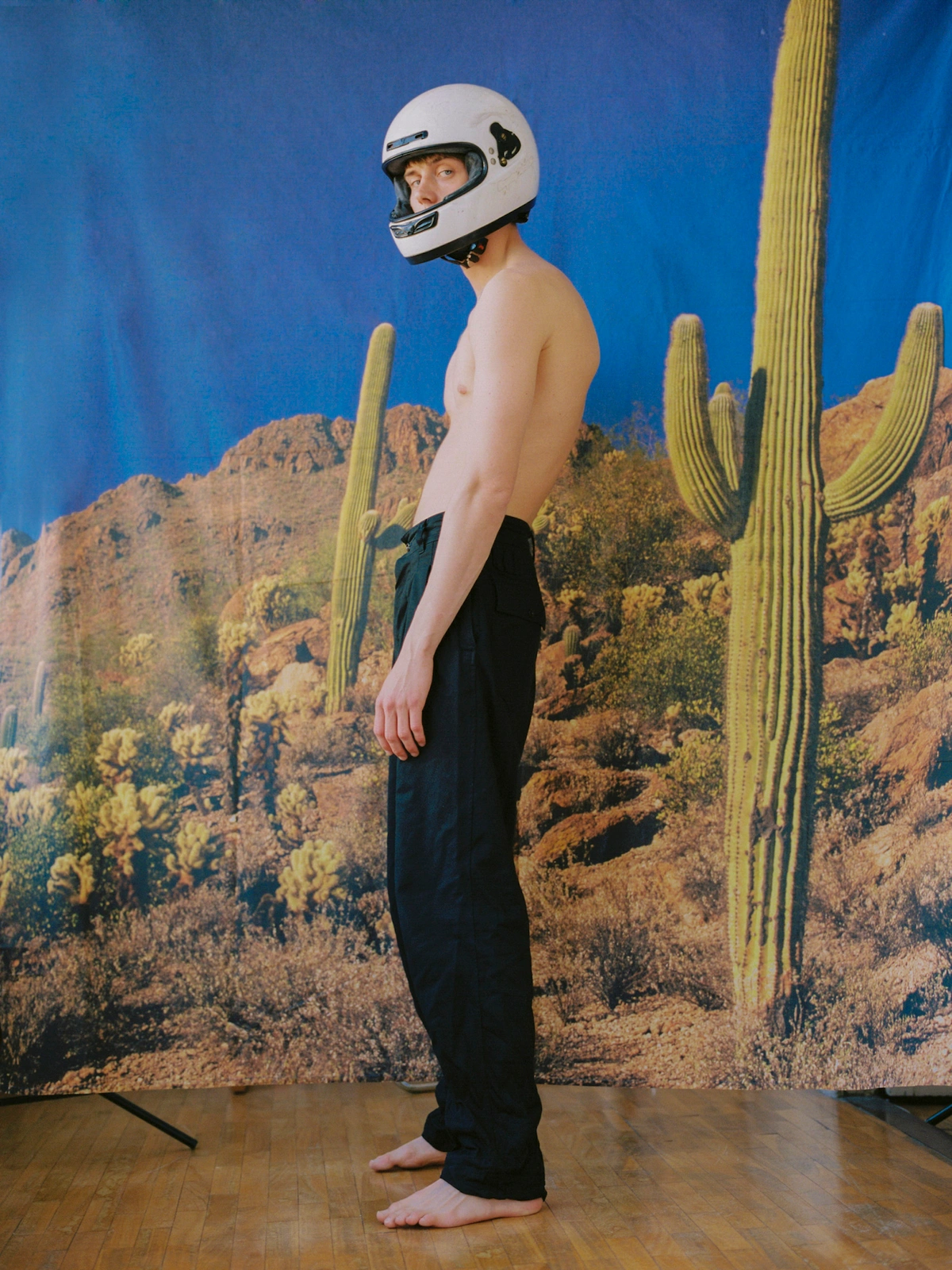 Shirtless man wearing a white motorcycle helmet and black trousers standing against a desert backdrop.