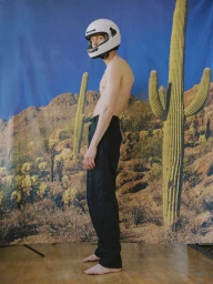Shirtless man wearing a white motorcycle helmet and black trousers standing against a desert backdrop.