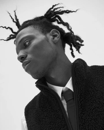 Black and white low-angle portrait of a Black man with dreadlocks wearing a sherpa fleece vest, white shirt, and tie against a plain backdrop.