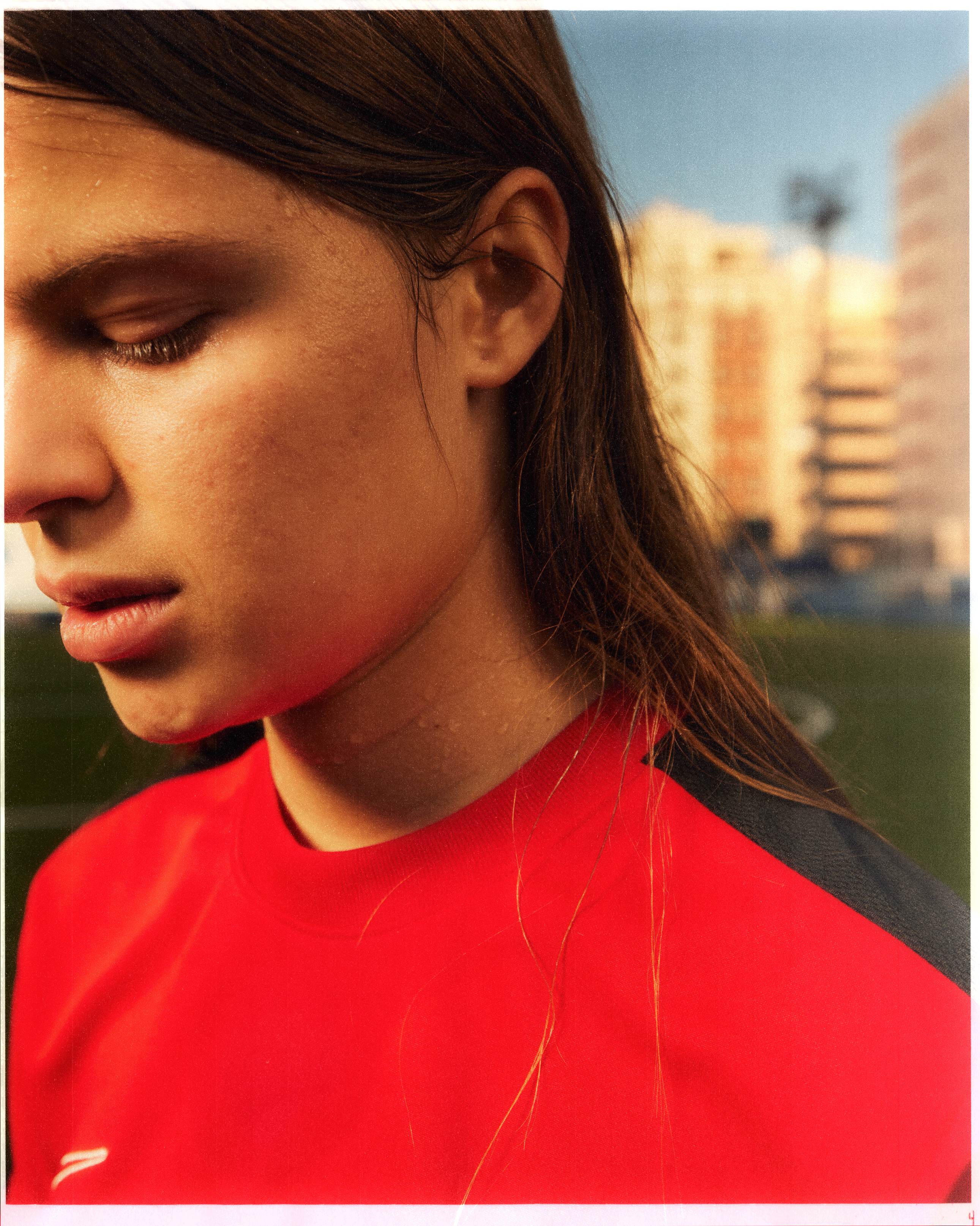 Close-up of a young adult with long brown hair wearing a red sport shirt with a neutral expression in an outdoor setting.