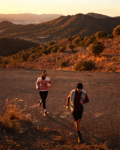 Two adults run uphill on a dirt path near mountains at sunset, wearing athletic apparel and hats, capturing a fitness scene.