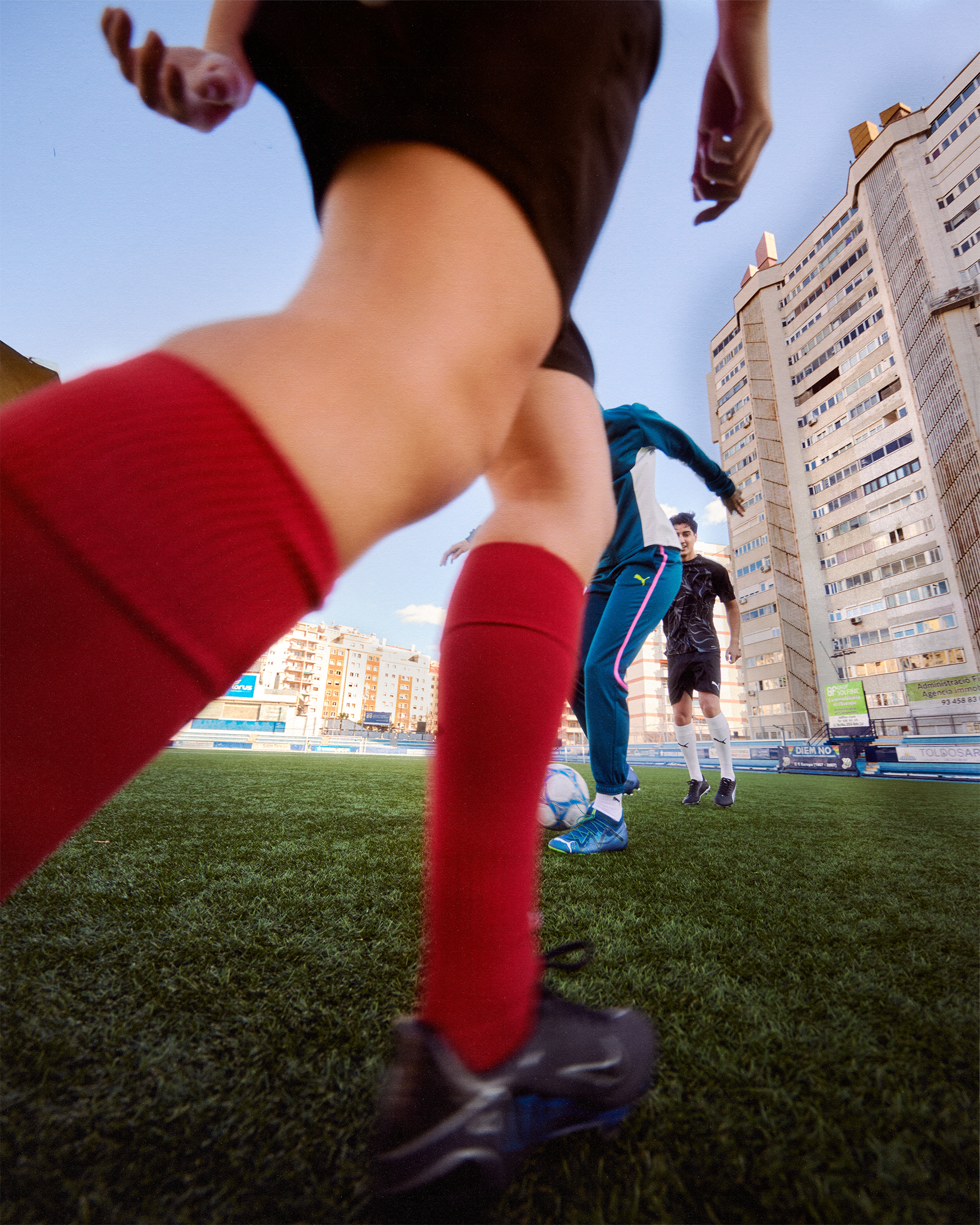 Close-up of soccer players in action on green turf field with urban building background, capturing the energy and focus of the game.