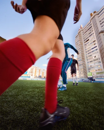 Close-up of soccer players in action on green turf field with urban building background, capturing the energy and focus of the game.