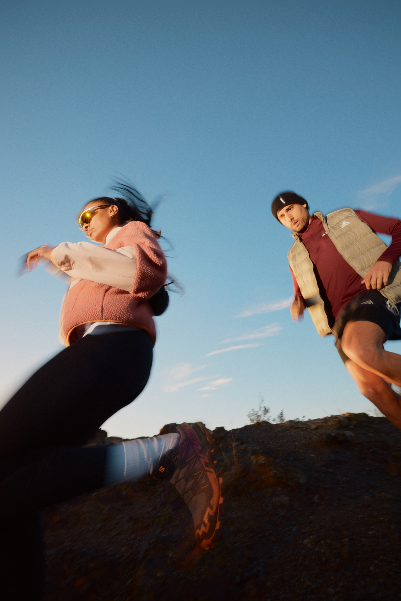 Two sports models are running outdoors on a trail with blue sky. One is wearing sunglasses, and the other is wearing a knit cap.