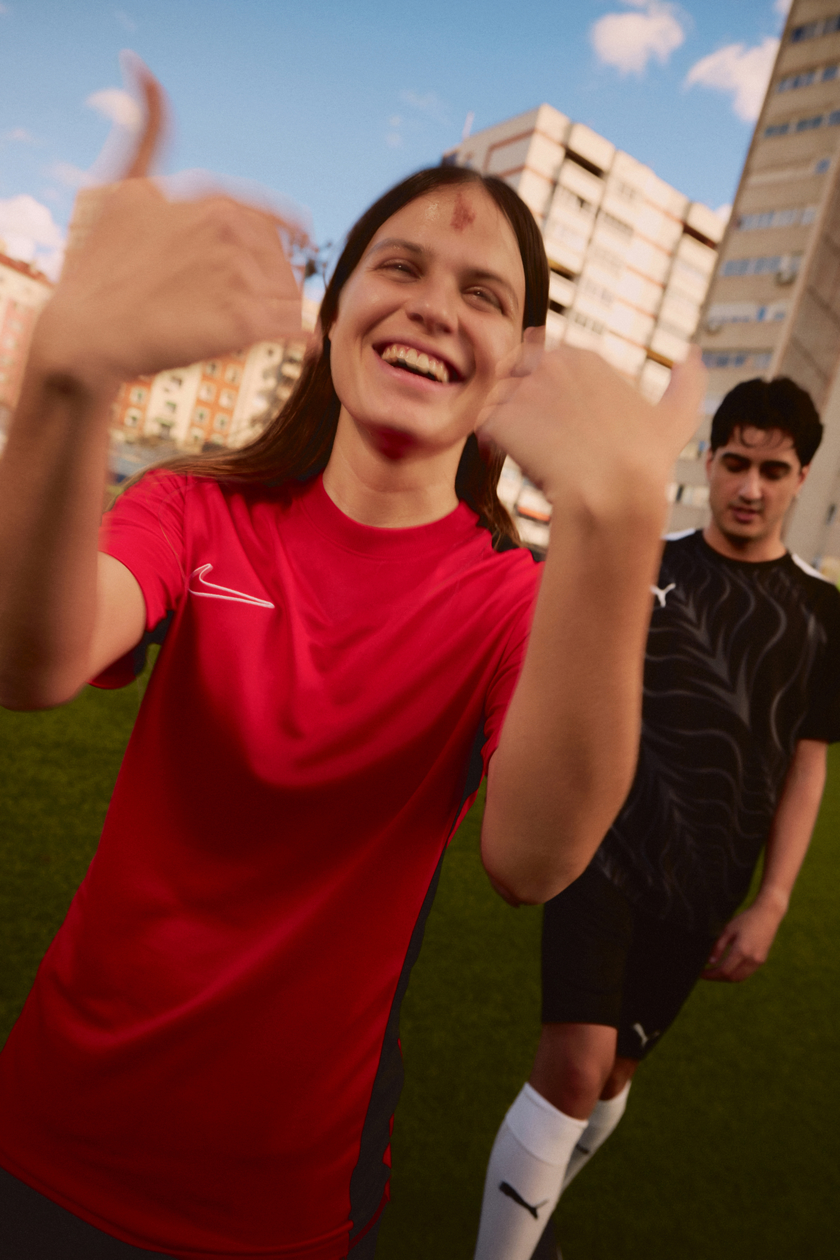 Two young adults smile and gesture in a medium shot. A woman in a red sports shirt is in the foreground, with an apartment block behind.