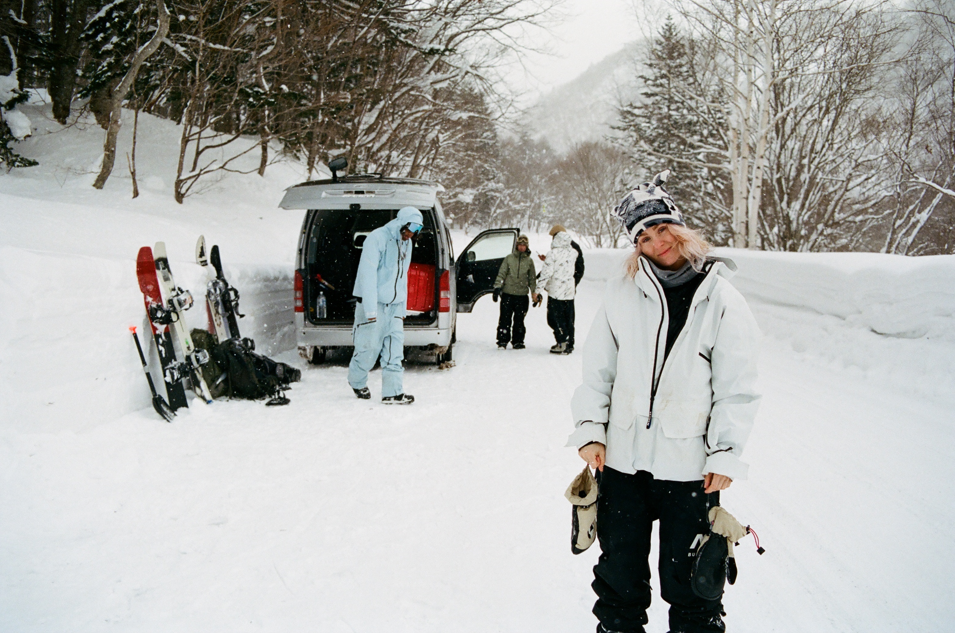 A woman in a white ski jacket and hat stands in snowy mountains, near a van with open doors and skis, creating a winter sport scene.