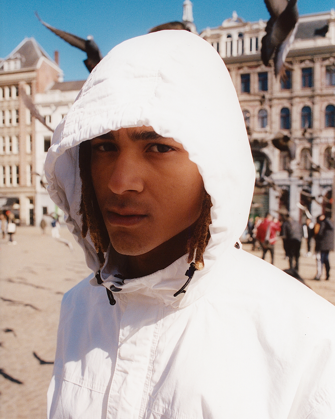 Man in white hooded jacket stands in a sunny urban square with pigeons flying around, set against historic buildings.