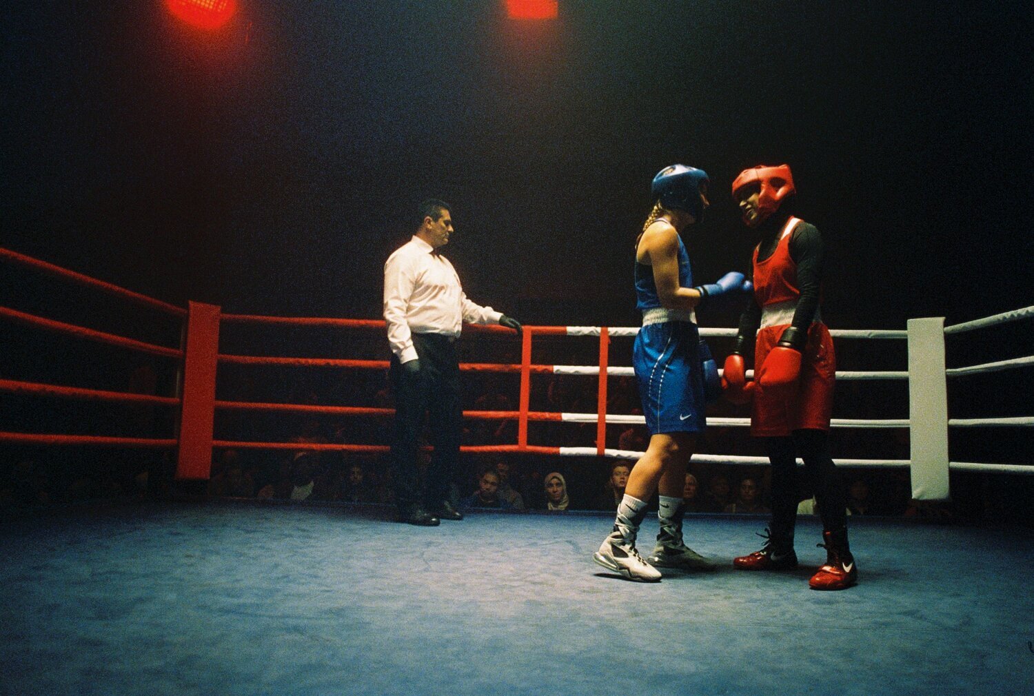 Two boxers face off in the boxing ring as the referee stands nearby, the crowd can be seen blurred in the background.