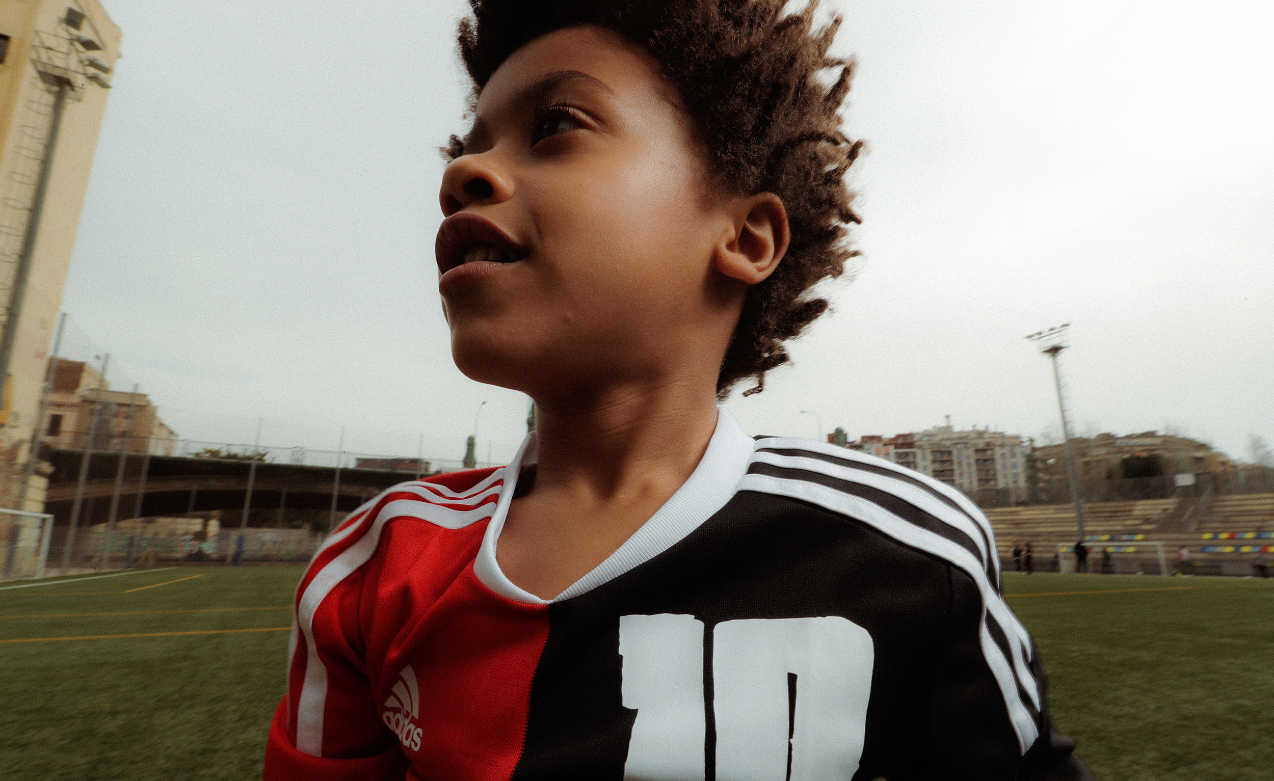 A young African-American boy in a red and black soccer jersey stands on a green soccer field on an overcast day, looking up and to the right.