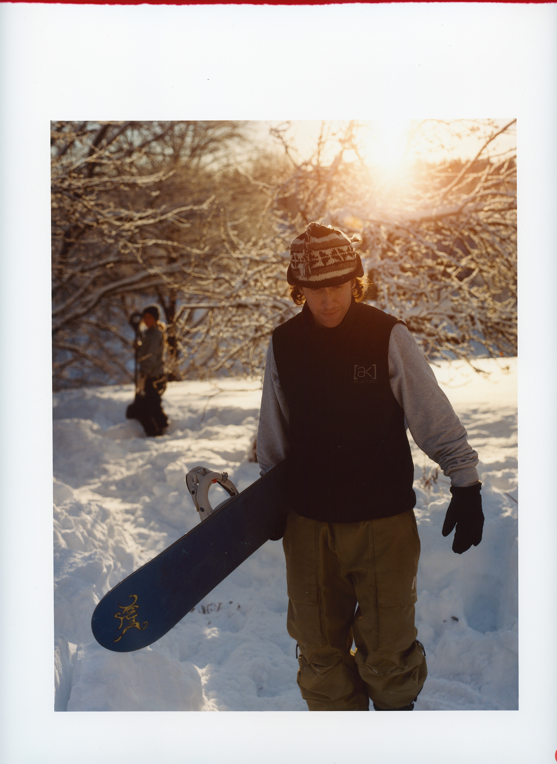 A male snowboarder in winter apparel walking in snowy landscape holding his snowboard, backlit by the sun with trees in background.