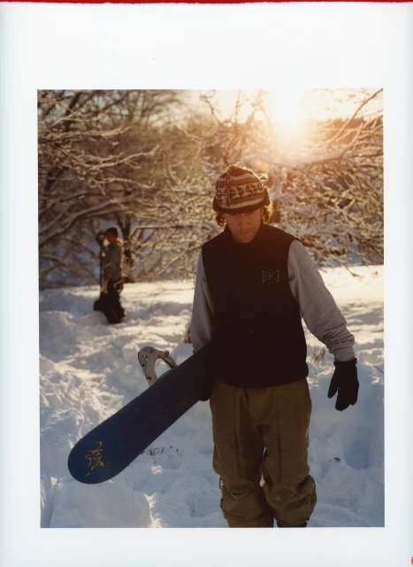 A male snowboarder in winter apparel walking in snowy landscape holding his snowboard, backlit by the sun with trees in background.