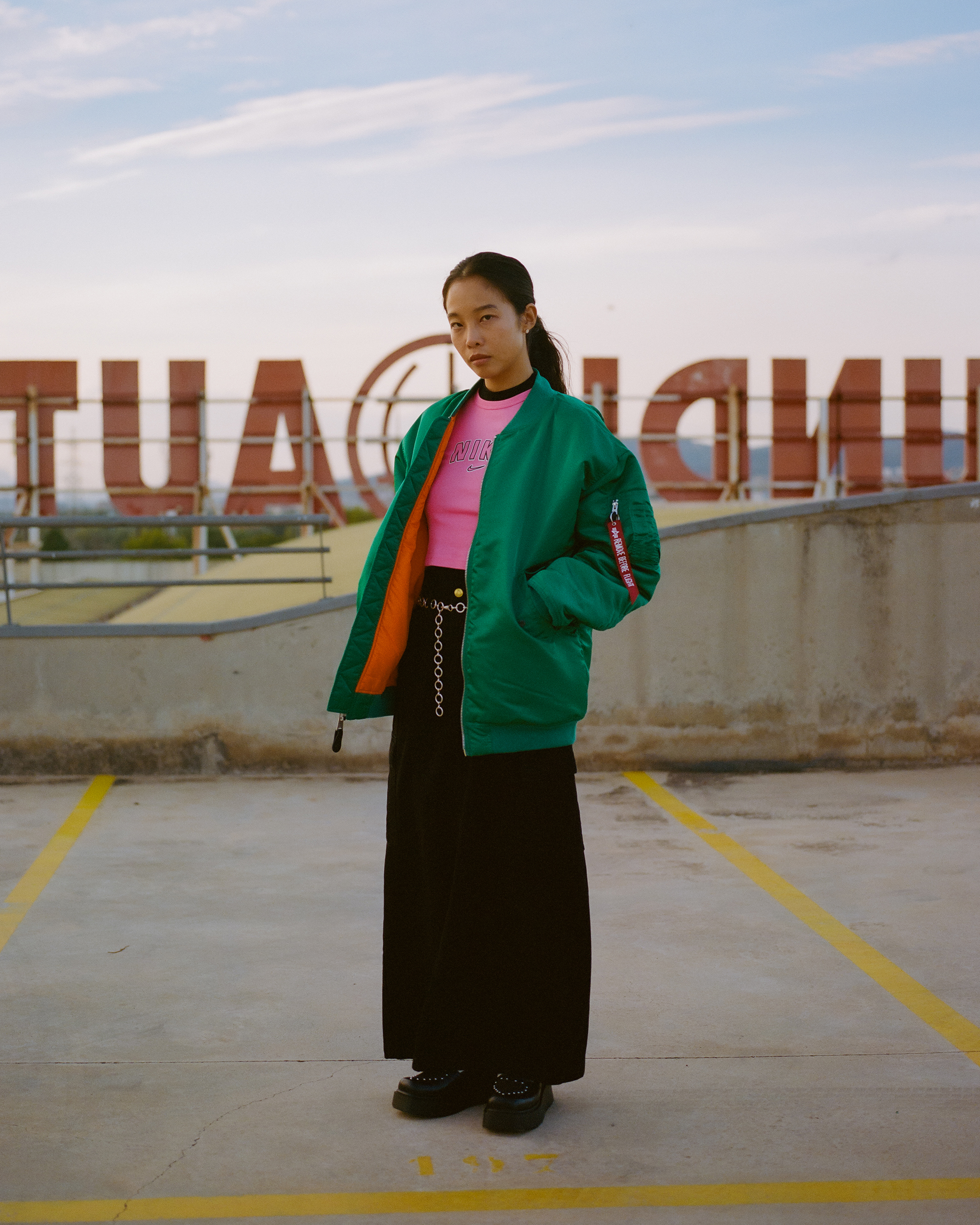 Young woman poses in a green bomber jacket and black skirt on a parking deck with an oversized sign in the background at dusk.