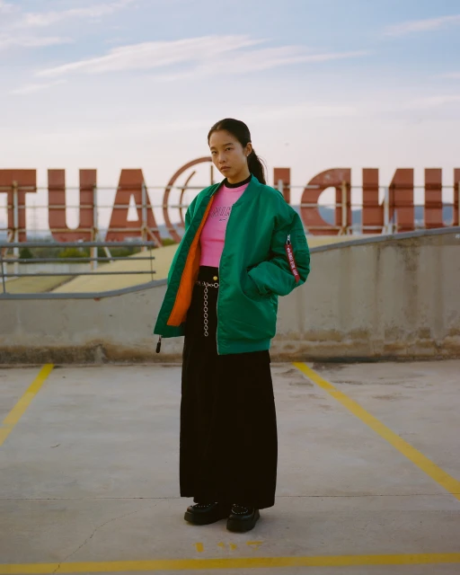Young woman poses in a green bomber jacket and black skirt on a parking deck with an oversized sign in the background at dusk.