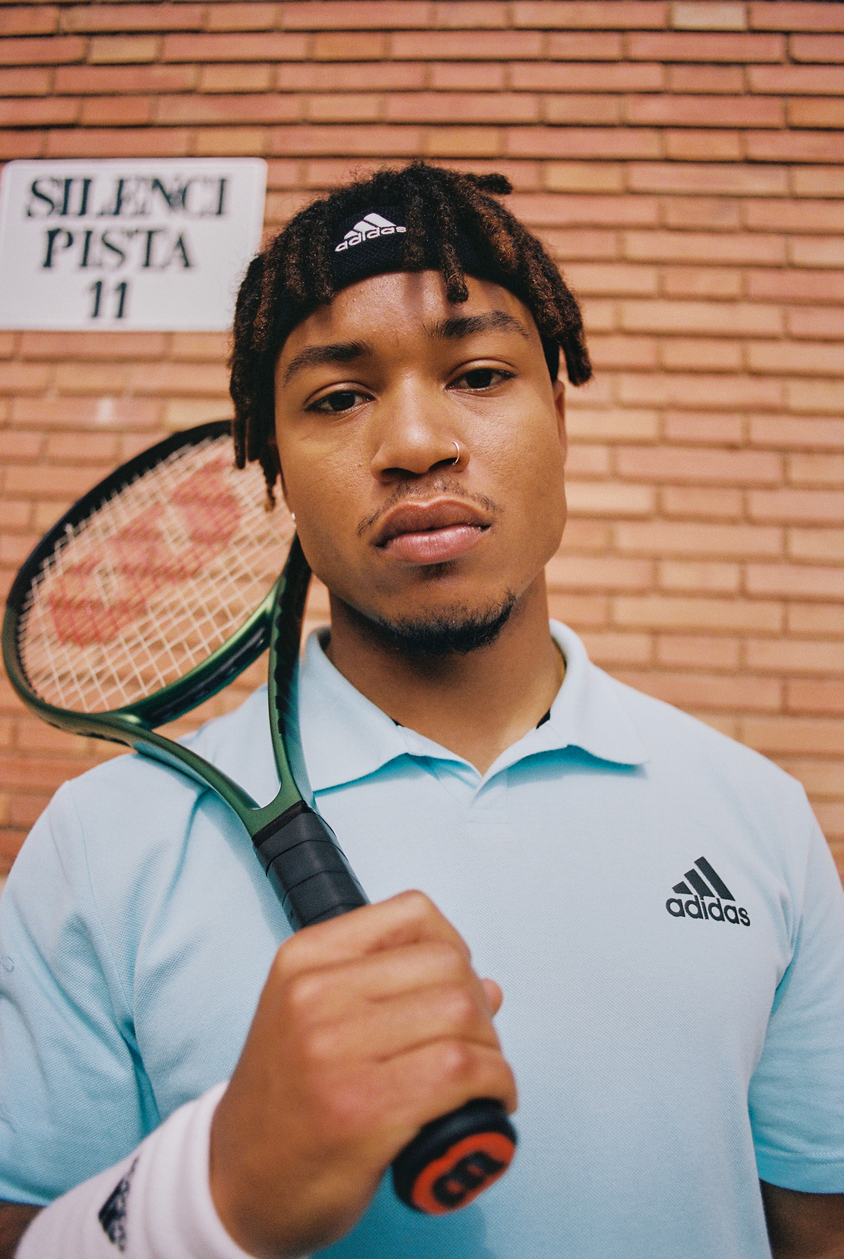 Portrait of a male tennis player with dreadlocks wearing an Adidas headband and shirt, holding a tennis racket, against a brick wall.