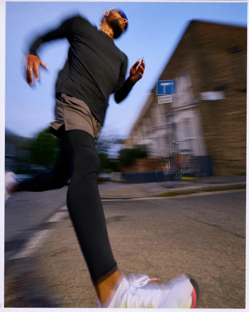 A man runs on a city street wearing athletic wear, creating a motion blur effect with a brick building and blue sign in the background.